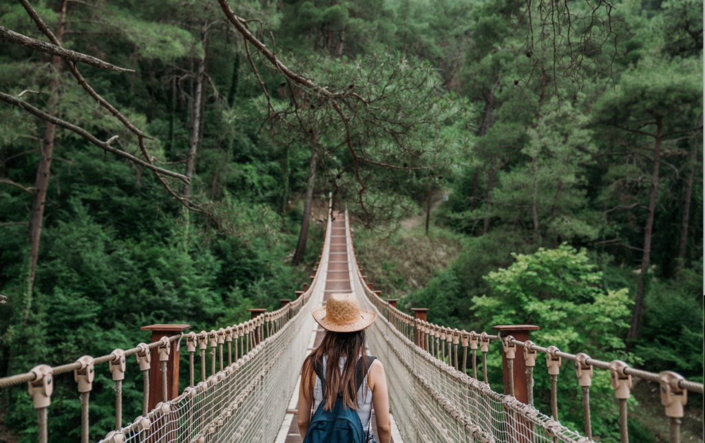  A woman wearing a hat and a backpack walks on a suspension bridge in a lush green forest.